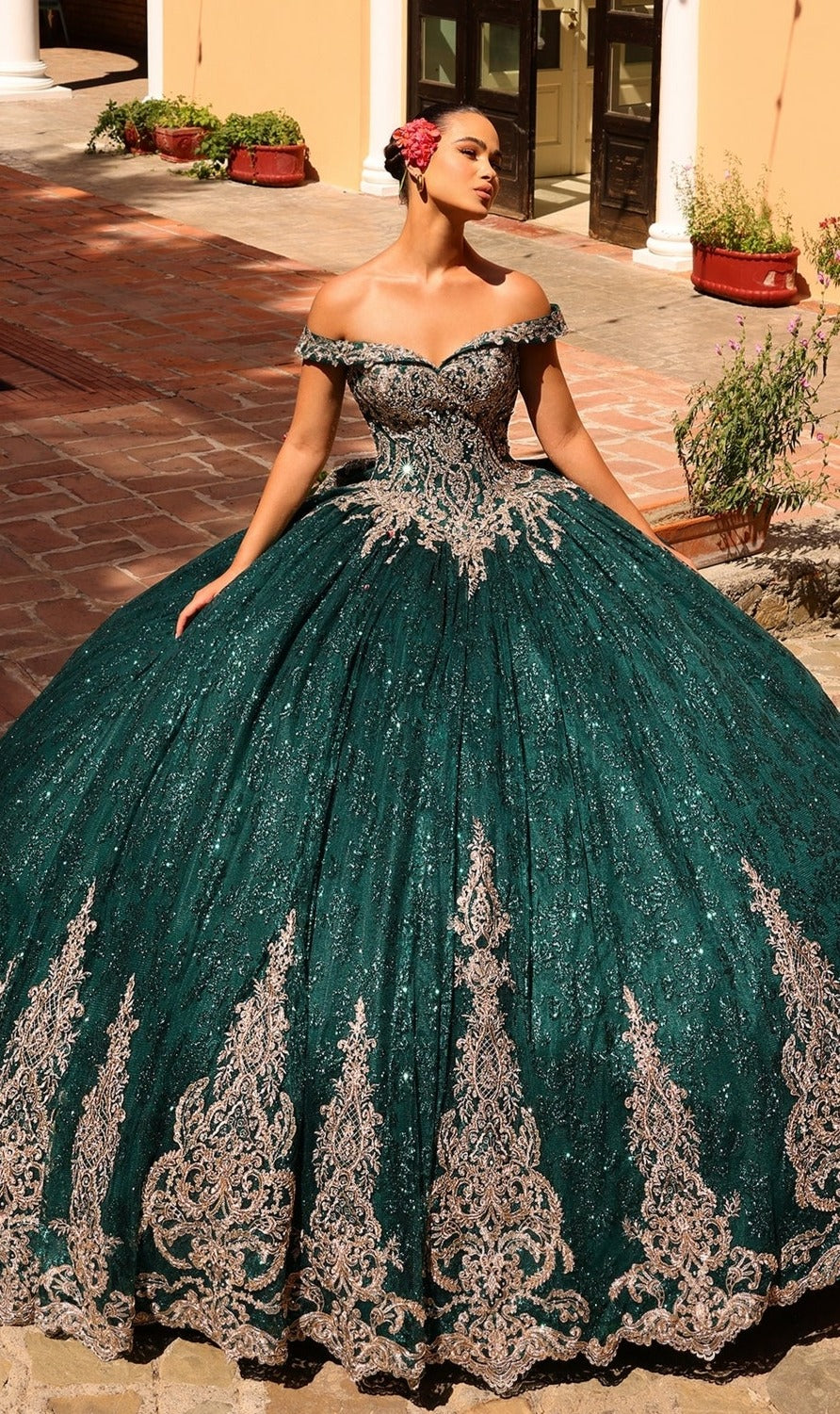 A woman poses outdoors in the Quinceanera Dress 54324 by Amarra—an off-the-shoulder, emerald green ball gown with intricate lace embroidery and a voluminous skirt.